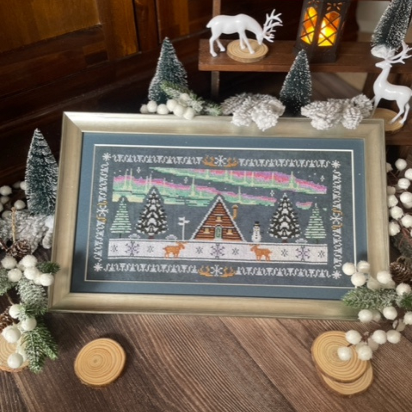 A stylised rectangular cross-stitch design depicting a cozy alpine chalet amid a snowy night scene. The central wooden chalet is flanked by tall, snow-covered evergreen trees. Above, a glowing aurora borealis arcs across the dark sky, rendered in soft flowing bands. Accented with swirling snowflakes or stars, the scene is framed by a decorative border that echoes the northern-lights palette.
