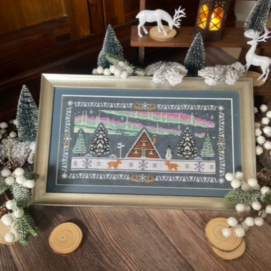 A stylised rectangular cross-stitch design depicting a cozy alpine chalet amid a snowy night scene. The central wooden chalet is flanked by tall, snow-covered evergreen trees. Above, a glowing aurora borealis arcs across the dark sky, rendered in soft flowing bands. Accented with swirling snowflakes or stars, the scene is framed by a decorative border that echoes the northern-lights palette.